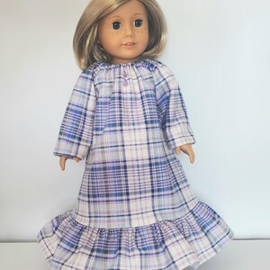 May include: A doll wearing a long-sleeved, plaid nightgown in shades of blue, purple, and white. The nightgown has a ruffled hem and a button closure at the neckline. The doll has blonde hair and is standing against a white background.
