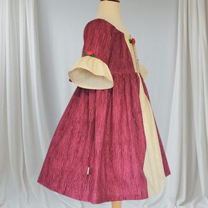 May include: A burgundy dress with a textured pattern, featuring cream-coloured accents and bell sleeves. The dress has a round neckline and is adorned with small red rose accents. The dress is displayed on a wooden mannequin.