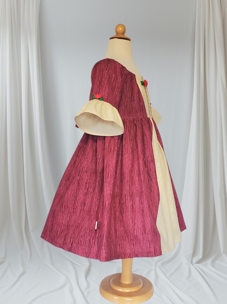 May include: A burgundy dress with a textured pattern, featuring cream-coloured accents and bell sleeves. The dress has a round neckline and is adorned with small red rose accents. The dress is displayed on a wooden mannequin.