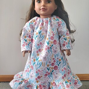 May include: A doll wearing a floral print nightgown. The white fabric is patterned with pink, yellow, and blue flowers and leaves. The nightgown has long sleeves and a ruffled hem. The doll has long brown hair and brown eyes.