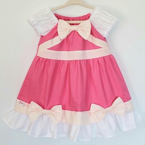 May include: A pink dress with white puff sleeves and a cream bow at the neckline. The dress has cream bows and trim along the hemline. The dress is hanging on a wooden hanger.