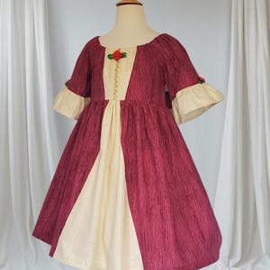 May include: A burgundy and cream-coloured dress with a textured fabric. The dress features a cream-coloured centre panel and sleeves with cream-coloured cuffs. The bodice is adorned with a red rose and gold trim. The dress is displayed on a wooden mannequin.