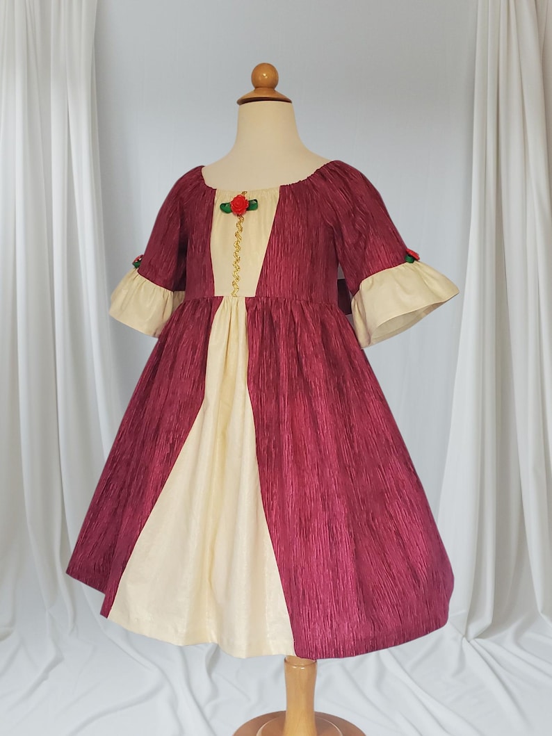 May include: A burgundy and cream-coloured dress with a textured fabric. The dress features a cream-coloured centre panel and sleeves with cream-coloured cuffs. The bodice is adorned with a red rose and gold trim. The dress is displayed on a wooden mannequin.
