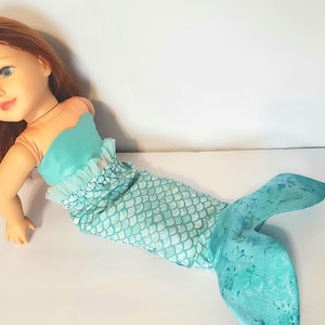 May include: A doll with red hair and blue eyes, dressed as a mermaid. The doll's top is teal, and the mermaid tail is teal with a scale pattern. The doll is lying on a white surface.