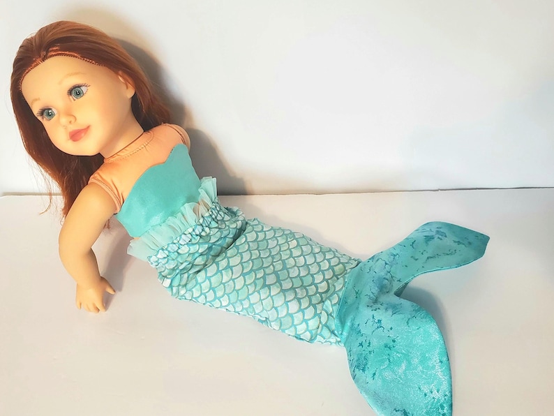 May include: A doll with red hair and blue eyes, dressed as a mermaid. The doll's top is teal, and the mermaid tail is teal with a scale pattern. The doll is lying on a white surface.