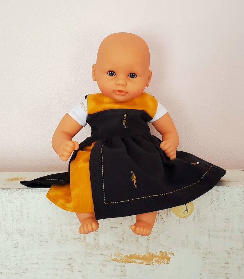 Puede incluir: Una mu&ntilde;eca calva con un vestido negro y dorado con abejas bordadas. La mu&ntilde;eca est&aacute; sentada sobre una superficie blanca.