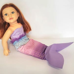 以下が含まれることがあります： 赤い髪と青い目の人魚の姿をした人形。人形のトップスはオレンジ、紫、青のグラデーションです。人魚の尾は金色の鱗模様のピンク色で、紫色の尾びれに変わります。