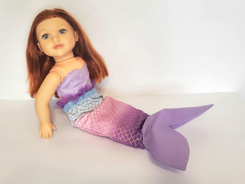 以下が含まれることがあります： 赤い髪と青い目の人魚の姿をした人形。人形のトップスはオレンジ、紫、青のグラデーションです。人魚の尾は金色の鱗模様のピンク色で、紫色の尾びれに変わります。
