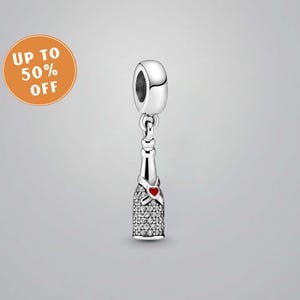 Peut inclure: Un charm argenté en forme de bouteille de champagne, orné de cristaux transparents et d'un cœur rouge. Le charm possède une boucle en haut pour l'attacher à un bracelet ou un collier. L'image comprend également un graphique orange circulaire avec le texte "UP TO 50% OFF".