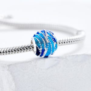 May include: A blue and silver bead charm with a wave-like design. The bead is a mix of dark and light blue, with silver accents and a silver core. The charm is on a silver bracelet.