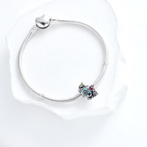 Puede incluir: Una pulsera plateada con un cierre en forma de coraz&oacute;n y un charm decorativo. El charm presenta un dise&ntilde;o colorido con detalles azules, rojos y verdes. La pulsera se muestra sobre una superficie blanca.
