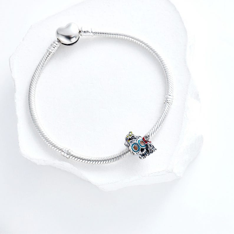 Puede incluir: Una pulsera plateada con un cierre en forma de coraz&oacute;n y un charm decorativo. El charm presenta un dise&ntilde;o colorido con detalles azules, rojos y verdes. La pulsera se muestra sobre una superficie blanca.
