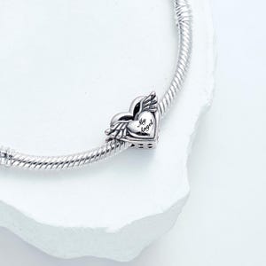Op de afbeelding: Een zilveren bedel met een hart- en vleugelontwerp, bevestigd aan een zilveren armband. Het hart heeft de tekst "My Angel" gegraveerd. De bedel is gedetailleerd en heeft een gepolijste afwerking.