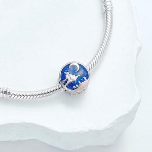 May include: A silver charm with a blue enamel design featuring a crescent moon and a silhouette of a person riding a flying carpet. The charm is attached to a silver bracelet, set against a white background.