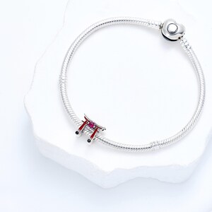 K&ouml;nnte beinhalten: Ein silbernes Charm-Armband mit einem japanischen Torii-Tor-Charm. Das Tor ist rot mit rosa Akzenten und schwarzen Details. Das Armband hat einen herzf&ouml;rmigen Verschluss und wird auf einer wei&szlig;en Oberfl&auml;che pr&auml;sentiert.