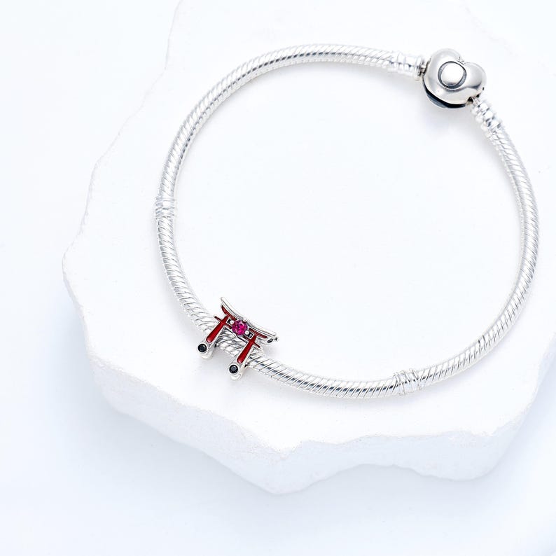 K&ouml;nnte beinhalten: Ein silbernes Charm-Armband mit einem japanischen Torii-Tor-Charm. Das Tor ist rot mit rosa Akzenten und schwarzen Details. Das Armband hat einen herzf&ouml;rmigen Verschluss und wird auf einer wei&szlig;en Oberfl&auml;che pr&auml;sentiert.