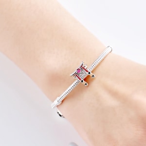 K&ouml;nnte beinhalten: Ein silbernes Armband mit einem Charm, das ein japanisches Torii-Tor darstellt. Der Charm ist mit roten und schwarzen Akzenten und einem kleinen, rosa Edelstein versehen. Das Armband hat ein strukturiertes Schlangenketten-Design.