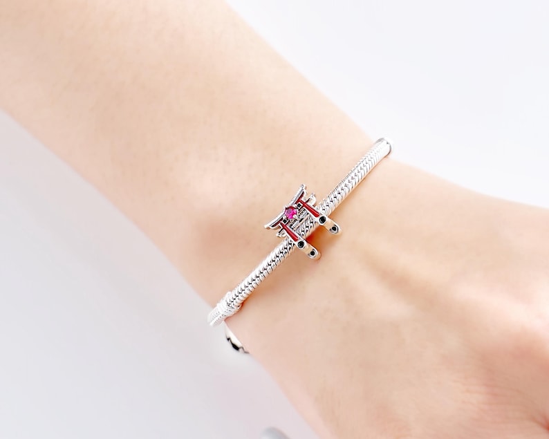K&ouml;nnte beinhalten: Ein silbernes Armband mit einem Charm, das ein japanisches Torii-Tor darstellt. Der Charm ist mit roten und schwarzen Akzenten und einem kleinen, rosa Edelstein versehen. Das Armband hat ein strukturiertes Schlangenketten-Design.