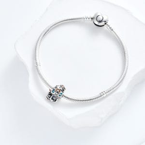 Puede incluir: Una pulsera de plata con un charm decorativo. El charm presenta un dise&ntilde;o colorido con detalles azules, naranjas y blancos. La pulsera tiene un cierre en forma de coraz&oacute;n.