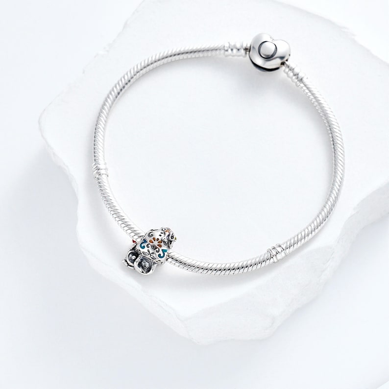 Puede incluir: Una pulsera de plata con un charm decorativo. El charm presenta un dise&ntilde;o colorido con detalles azules, naranjas y blancos. La pulsera tiene un cierre en forma de coraz&oacute;n.