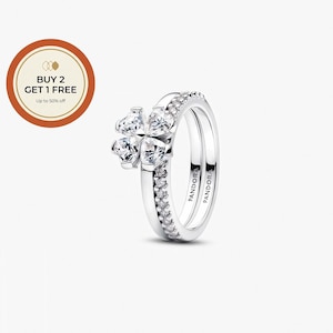 Op de afbeelding: Twee zilveren ringen. Eén ring heeft een klaverbladontwerp met heldere, sprankelende stenen. De tweede ring is een band met kleine, heldere stenen. Het woord "PANDORA" is aan de binnenkant van de ringen gegraveerd.