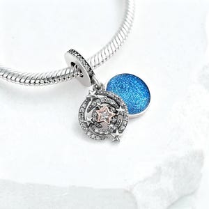 May include: A silver charm with a celestial theme. The charm features a circular design with embedded clear crystals and star accents, surrounding a central rose gold star. A separate blue, glitter-filled disc hangs nearby.