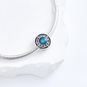 May include: A silver charm bracelet with a round, decorative charm. The charm features a blue centre with a geometric design in black and silver. The bracelet is on a white surface.