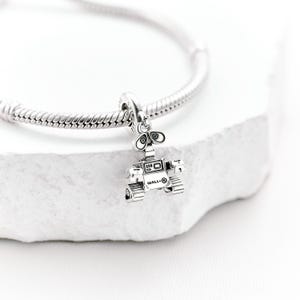 May include: A silver charm depicting the character Wall-E from the animated film. The charm is attached to a silver bracelet. The robot charm has a boxy body, two large eyes, and tank treads. The word "WALL-E" is visible on the charm.