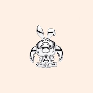 May include: A silver-colored charm in the shape of a cartoon bunny with long ears, arms, and a seated pose. The charm has a shiny, reflective surface and is set against a light peach background. The word "PANDORA" is visible on the charm.