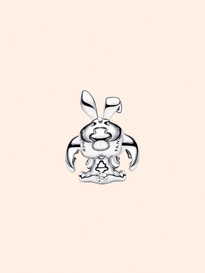 May include: A silver-colored charm in the shape of a cartoon bunny with long ears, arms, and a seated pose. The charm has a shiny, reflective surface and is set against a light peach background. The word "PANDORA" is visible on the charm.