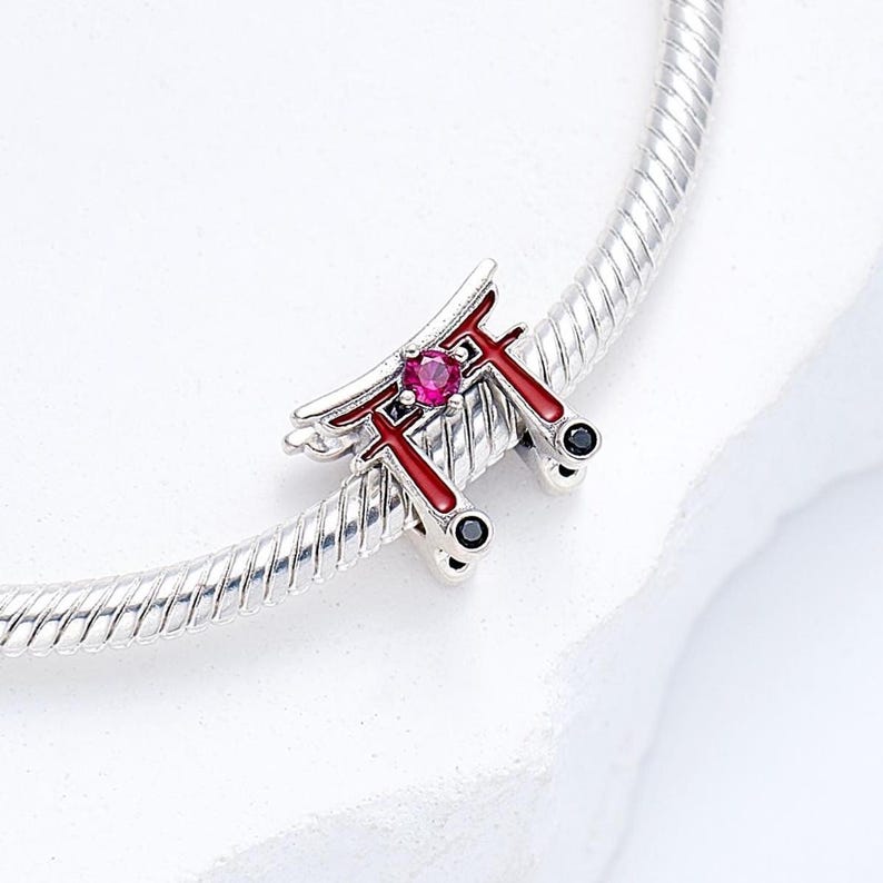 K&ouml;nnte beinhalten: Ein silberner Charm mit einem roten und silbernen Torii-Tor-Design, verziert mit einem rosa Edelstein und schwarzen Details. Der Charm ist an einem silbernen Armband befestigt, mit japanischem Thema.