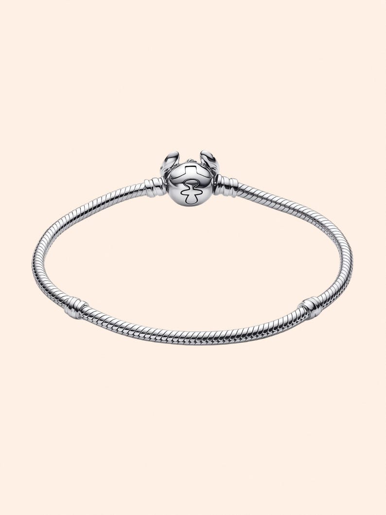 Puede incluir: Una pulsera plateada con un dije redondo con un dise&ntilde;o estilizado. La pulsera tiene un dise&ntilde;o de cadena de serpiente y un cierre de broche. El dije tiene un patr&oacute;n detallado y est&aacute; centrado en la pulsera.