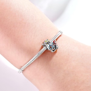 Puede incluir: Una pulsera de plata con una cuenta decorativa de calavera. La cuenta de calavera presenta acentos coloridos de rojo, verde y azul. La pulsera se lleva en la mu&ntilde;eca de una persona.