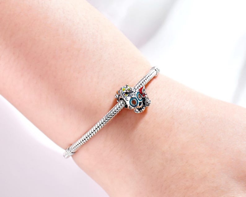 Puede incluir: Una pulsera de plata con una cuenta decorativa de calavera. La cuenta de calavera presenta acentos coloridos de rojo, verde y azul. La pulsera se lleva en la mu&ntilde;eca de una persona.
