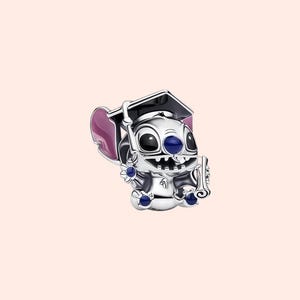 May include: A silver charm depicting a cartoon character wearing a graduation cap and holding a diploma. The charm has blue and purple accents and is set against a light pink background. The character has large black eyes and a wide smile.