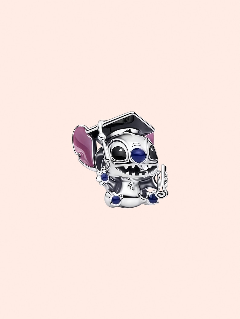 May include: A silver charm depicting a cartoon character wearing a graduation cap and holding a diploma. The charm has blue and purple accents and is set against a light pink background. The character has large black eyes and a wide smile.