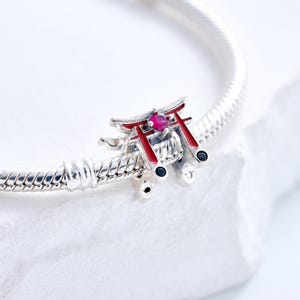 K&ouml;nnte beinhalten: Ein silberner Charm-Anh&auml;nger mit einem Miniatur-Torii-Tor aus Rot und Silber mit einem zentralen rosa Edelstein. Der Charm ist an einem silbernen Armband befestigt, vor einem wei&szlig;en Hintergrund. Das Torii-Tor-Design ist ein Symbol der japanischen Kultur.