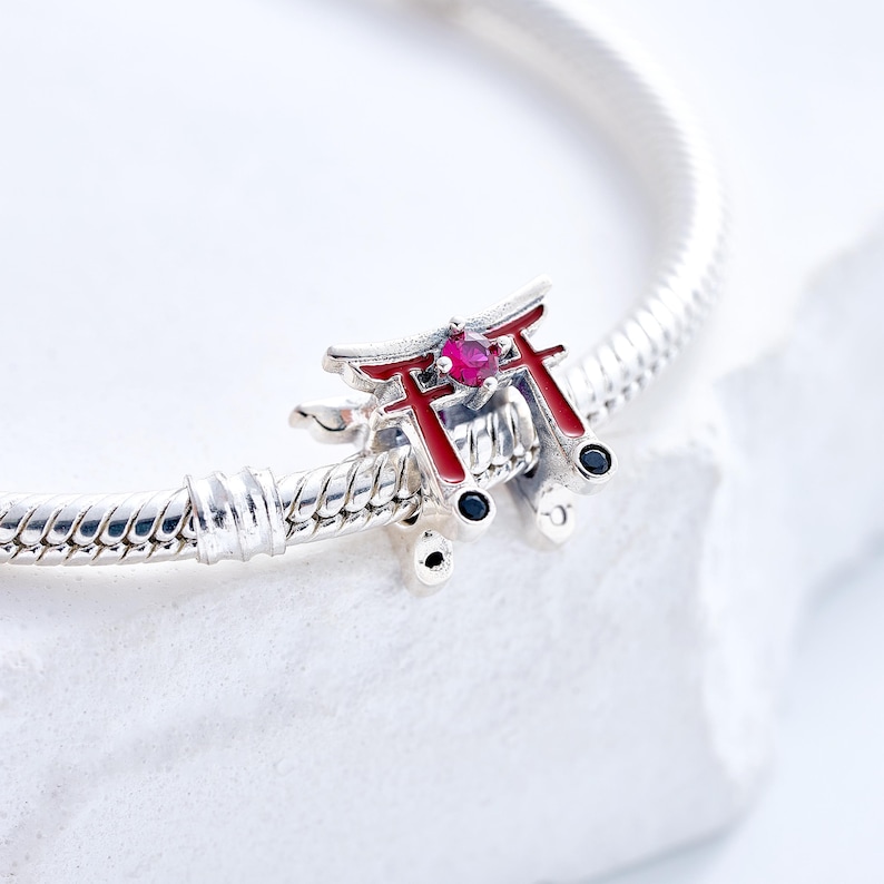 K&ouml;nnte beinhalten: Ein silberner Charm-Anh&auml;nger mit einem Miniatur-Torii-Tor aus Rot und Silber mit einem zentralen rosa Edelstein. Der Charm ist an einem silbernen Armband befestigt, vor einem wei&szlig;en Hintergrund. Das Torii-Tor-Design ist ein Symbol der japanischen Kultur.