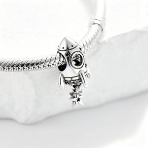 Könnte beinhalten: Ein silberner Charm-Anhänger in Form einer Rakete mit Sternaussparungen und einem Sternencluster. Der Charm ist an einem silbernen Armband befestigt, passend zum Weltraum-Thema.
