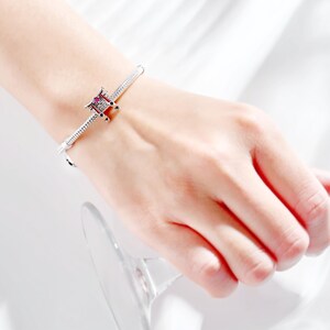 K&ouml;nnte beinhalten: Ein silbernes Armband mit einem Anh&auml;nger mit einem roten und schwarzen Design. Das Armband wird am Handgelenk getragen, mit einem Glas im Hintergrund. Der Anh&auml;nger hat ein detailliertes architektonisches Design.