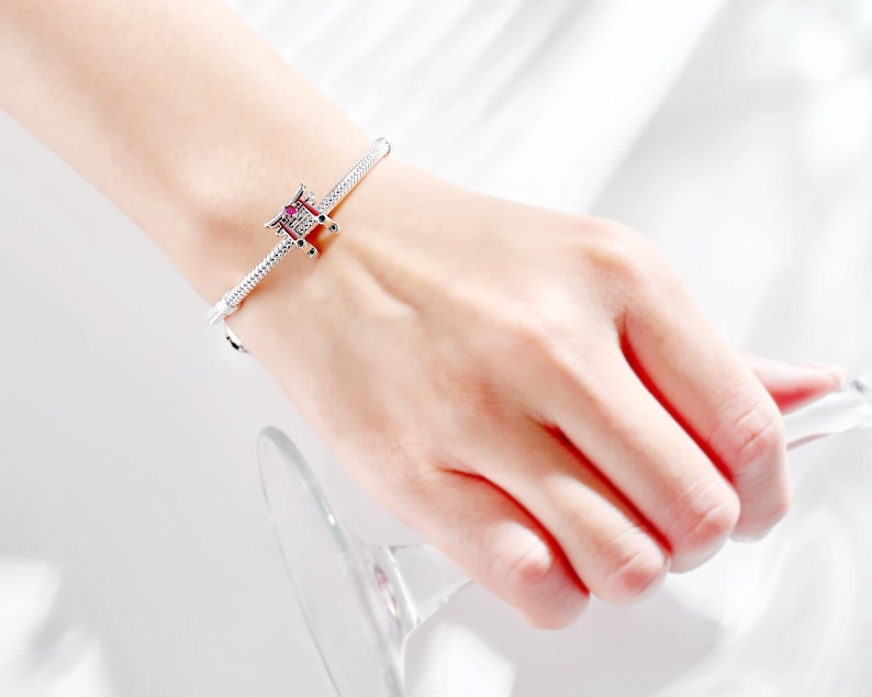 K&ouml;nnte beinhalten: Ein silbernes Armband mit einem Anh&auml;nger mit einem roten und schwarzen Design. Das Armband wird am Handgelenk getragen, mit einem Glas im Hintergrund. Der Anh&auml;nger hat ein detailliertes architektonisches Design.