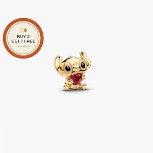 K&ouml;nnte beinhalten: Ein goldfarbener Charm-Anh&auml;nger mit der Figur Stitch aus Lilo & Stitch, der ein rotes herzf&ouml;rmiges Juwel h&auml;lt. Der Charm hat schwarze Augen und ist vor einem wei&szlig;en Hintergrund platziert. Der Charm ist ein Sammlerst&uuml;ck.