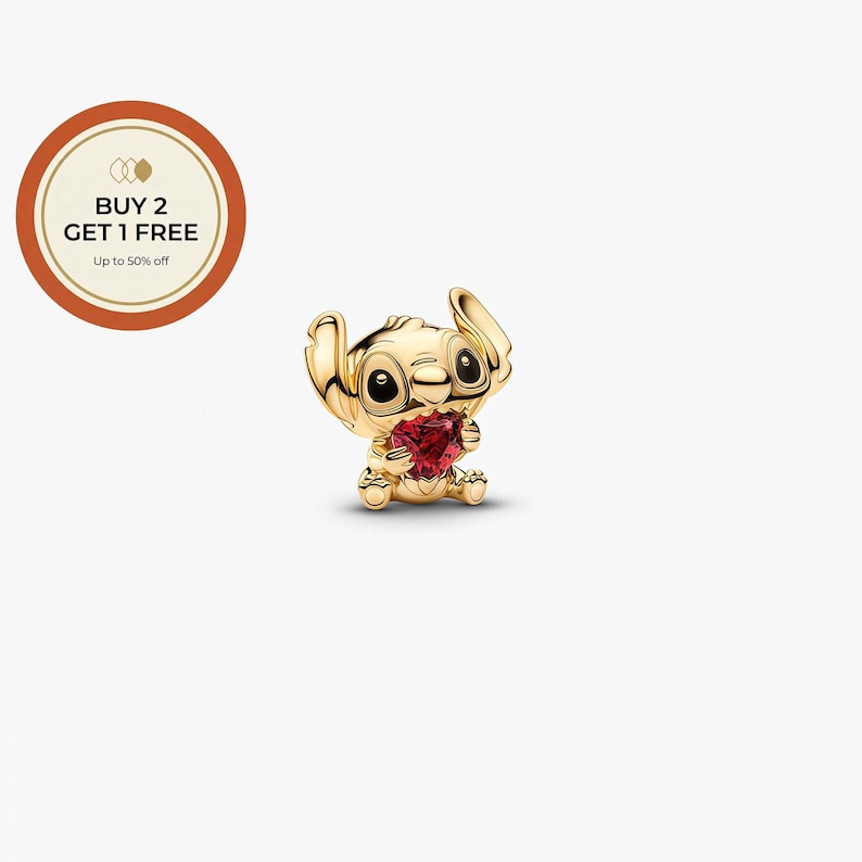 K&ouml;nnte beinhalten: Ein goldfarbener Charm-Anh&auml;nger mit der Figur Stitch aus Lilo & Stitch, der ein rotes herzf&ouml;rmiges Juwel h&auml;lt. Der Charm hat schwarze Augen und ist vor einem wei&szlig;en Hintergrund platziert. Der Charm ist ein Sammlerst&uuml;ck.