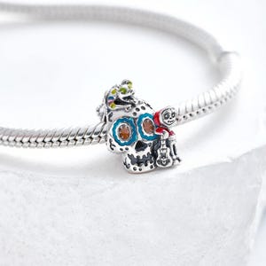 Puede incluir: Un charm de plata con un dise&ntilde;o detallado de calavera de az&uacute;car con detalles azules, naranjas y blancos. Una peque&ntilde;a figura con una capa roja con capucha y una guitarra est&aacute; unida a la calavera. Una peque&ntilde;a criatura verde y amarilla est&aacute; encima de la calavera.
