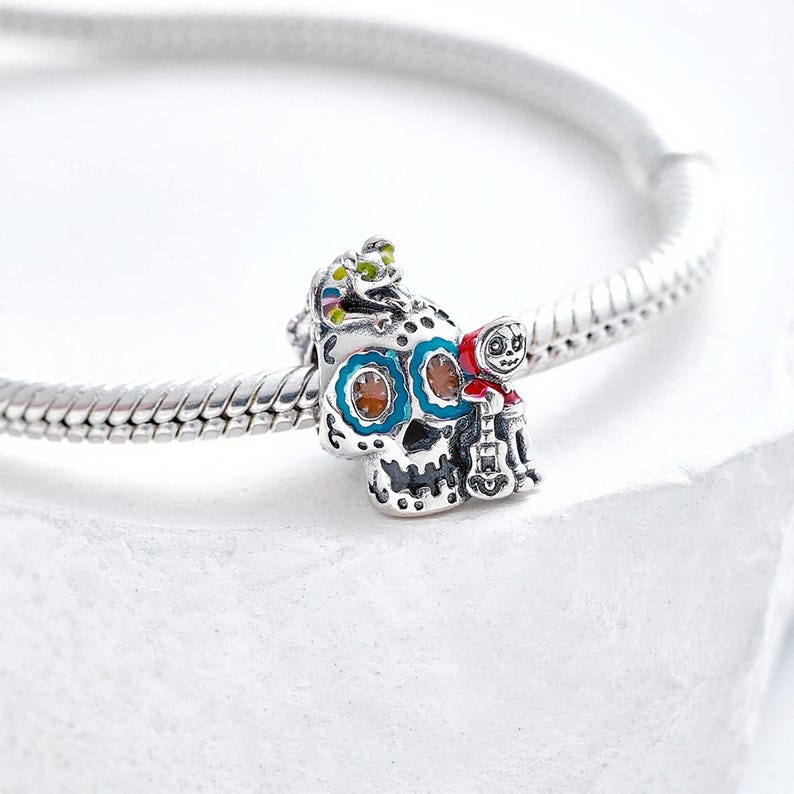 Puede incluir: Un charm de plata con un dise&ntilde;o detallado de calavera de az&uacute;car con detalles azules, naranjas y blancos. Una peque&ntilde;a figura con una capa roja con capucha y una guitarra est&aacute; unida a la calavera. Una peque&ntilde;a criatura verde y amarilla est&aacute; encima de la calavera.