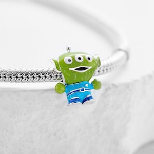 May include: A green and blue charm featuring a cartoon alien character with three eyes and a smiling mouth. The charm is attached to a silver bracelet. The alien is wearing a blue uniform with a belt and blue shoes. The charm is detailed and colorful.