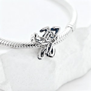 May include: A silver charm with an abstract design, possibly a stylized flower or knot, is attached to a silver bracelet. The charm has intricate details and a polished finish, reflecting light. The bracelet has a snake chain design.