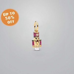 May include: A gold-coloured charm shaped like a toy soldier, with a red and gold hat and uniform. The charm features a small star and a clear gem on the hat. This festive charm is a holiday item.