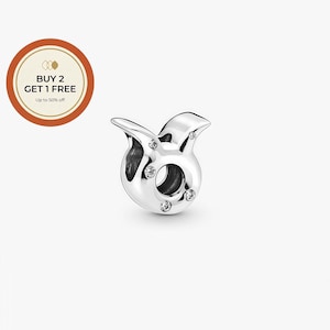 May include: A silver-colored charm in the shape of the Taurus zodiac symbol, featuring a polished finish and embedded with small, clear crystals. The charm has a circular opening in the center and two curved horns. The charm is on a white background.