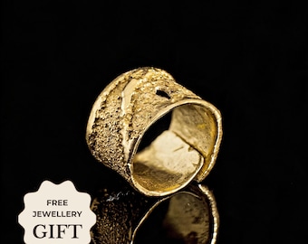 May include: A wide gold ring with a textured, uneven surface and a rough, organic finish. The band features an open design with irregular edges. A white badge in the corner displays the text FREE JEWELLERY GIFT in black capital letters.